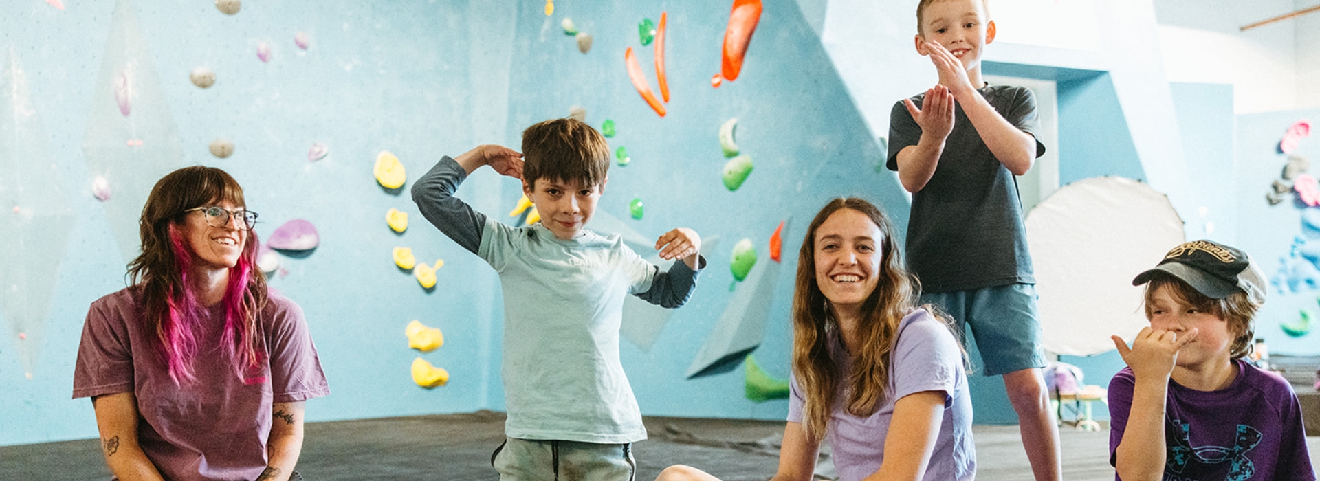 Four children and an adult pose cheerfully in a brightly colored indoor climbing gym, with holds on the walls behind them. It’s a perfect scene from Parents Night Out—everyone looks happy and relaxed.