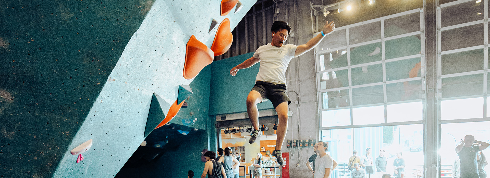 A man in a white t-shirt and shorts is falling or jumping off an indoor climbing wall with orange holds, mid-air, in a busy climbing gym with people watching—perfect for those enjoying their summer membership.