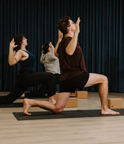 Three people practice yoga indoors at a climbing gym in Seattle’s University District, kneeling on mats with arms raised and bent. A dark curtain hangs in the background and yoga blocks sit beside them.