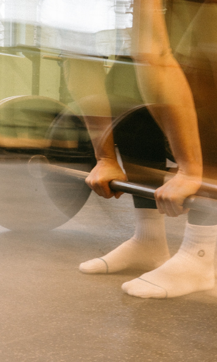 A person wearing white socks prepares to lift a barbell in a gym, their hands gripping the bar. The image is slightly blurred, suggesting motion—perfect for highlighting our flexible membership options and competitive pricing.
