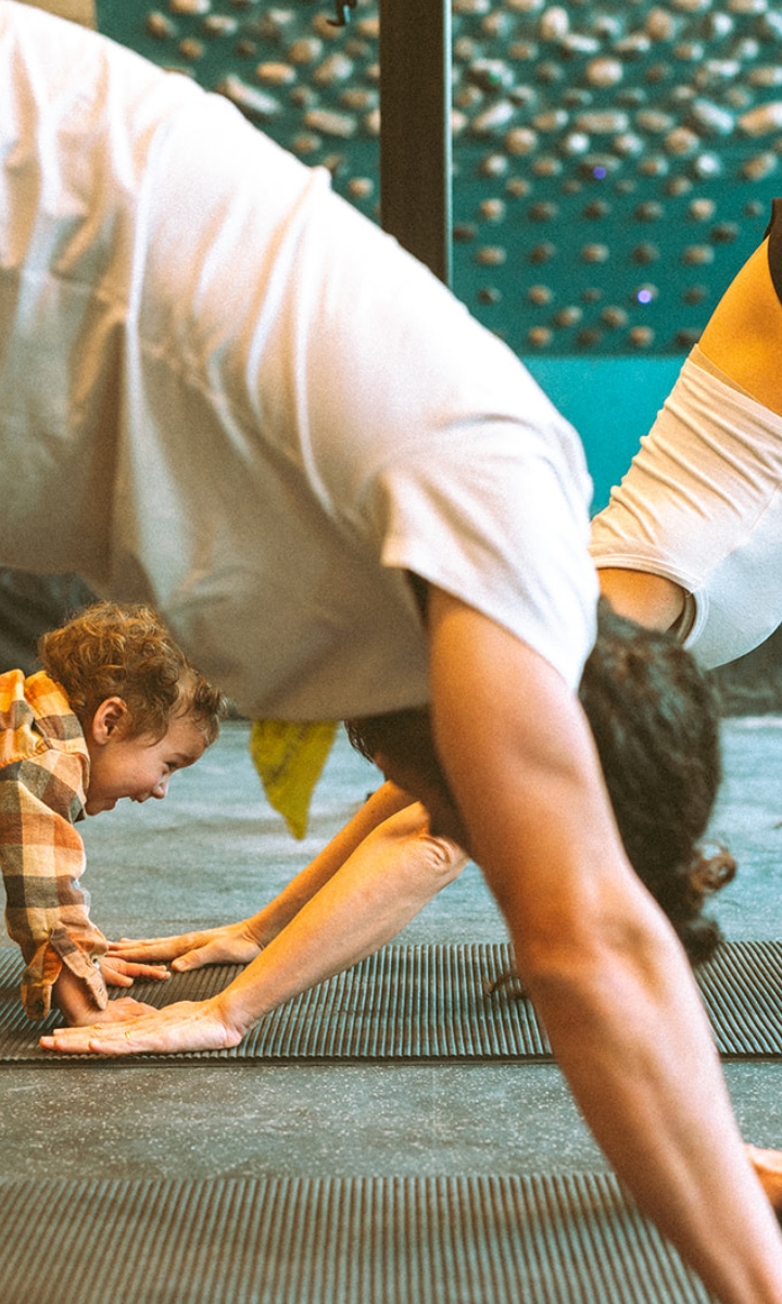 A young child mimics a person doing a downward dog yoga pose on a mat indoors, smiling and reaching forward while others practice beside them, experiencing the fun and benefits included with membership.