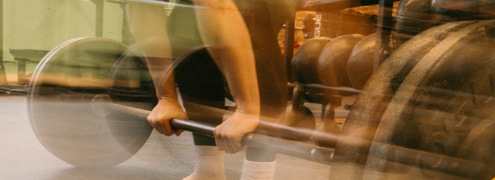 Person gripping a barbell in a gym, preparing to lift weights. The image has a motion blur effect, emphasizing movement and effort—perfect for showcasing membership benefits and competitive pricing. Gym equipment is visible in the background.