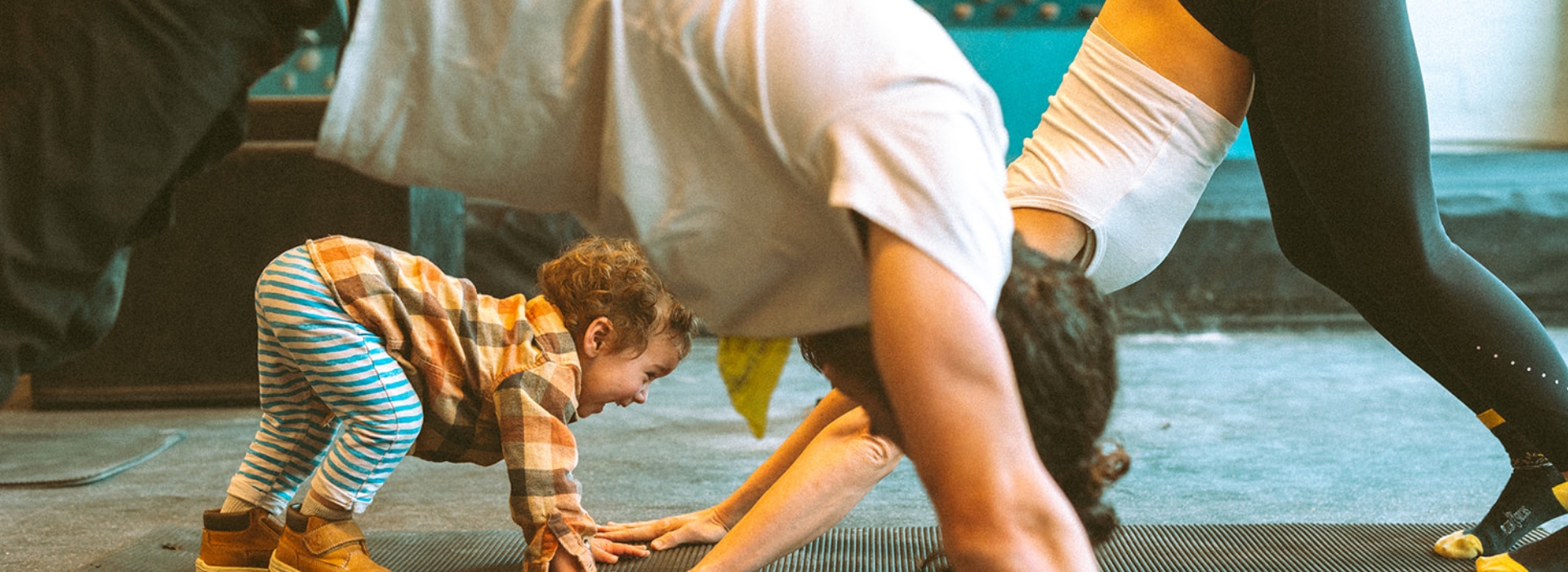 A toddler mimics adults doing yoga, playfully crawling under a persons arm while two adults hold downward dog poses on yoga mats indoors—showcasing the playful side of family yoga at any membership or pricing level.