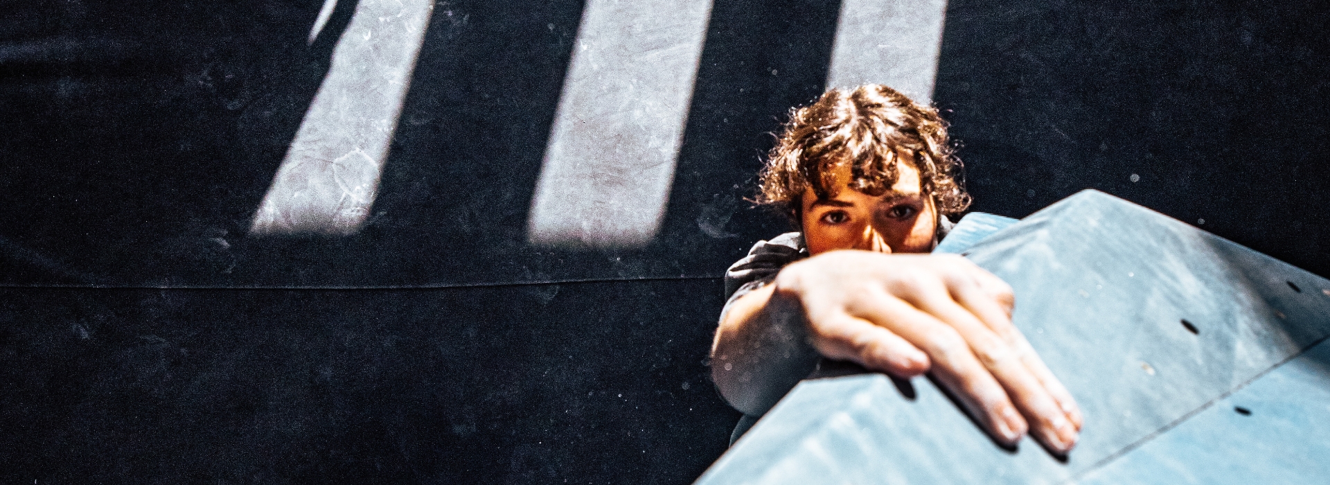 A person with curly hair reaches up and grasps a blue climbing hold, looking focused, with dramatic shadows and sunlight in the background—capturing the thrill of every climb included in your membership.