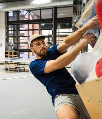 A man wearing glasses, a cap, and a navy shirt is climbing an indoor bouldering wall, gripping large handholds with focus. Tables and large windows are visible in the background.