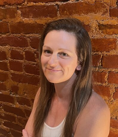 A woman with long brown hair and a white sleeveless top smiles softly while sitting in front of an orange-red brick wall, suggesting a cozy space perfect for relaxing after climbing. Warm indoor lighting highlights her face and the textured bricks behind her.