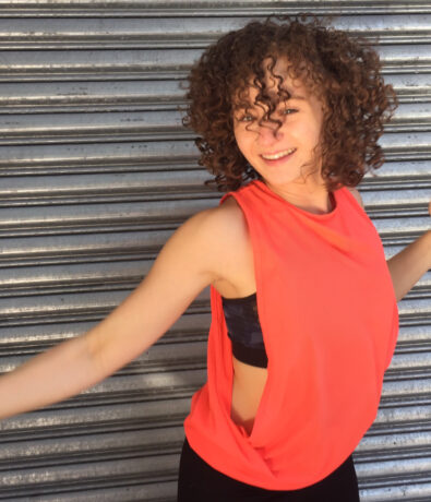 A smiling person with curly hair wearing a sleeveless bright orange top poses energetically in front of a metallic, corrugated background, as if ready for their next climbing adventure.