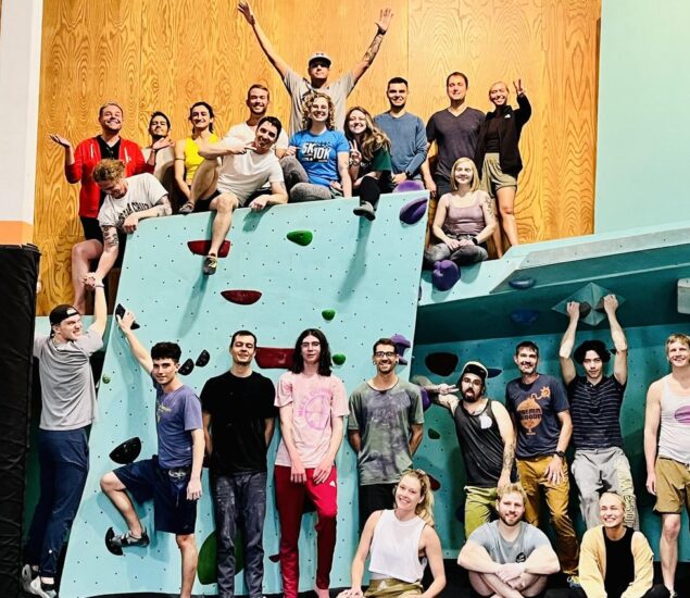 A large group of people pose and smile on an indoor climbing wall in Minneapolis. Some stand or sit on the wall, while others hang from holds—showcasing the vibrant spirit of local Community Clubs in a bright, lively gym setting.