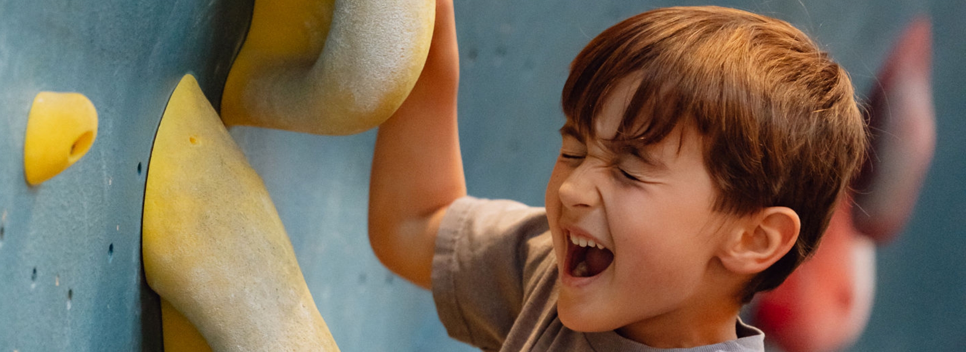 During the school holidays in Brooklyn, a young boy with brown hair climbs an indoor rock wall, eyes closed and mouth open in excitement or concentration, gripping a yellow climbing hold with one hand.