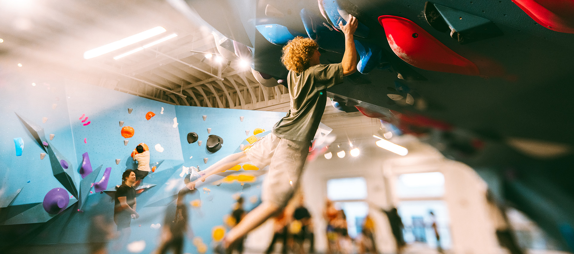 A person in shorts and a t-shirt climbs an indoor bouldering wall at Climbing Tempe, gripping holds with both hands. Other people are visible climbing and watching in the background of the brightly-lit gym.