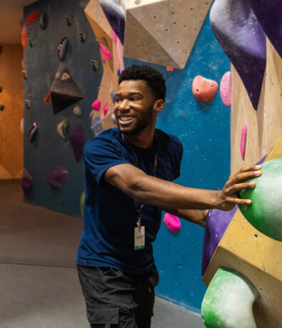 A man smiling and holding onto a climbing wall indoors, wearing a navy blue shirt and black shorts, with a staff badge around his neck. Colorful climbing holds highlight the indoor climbing environment in the background.