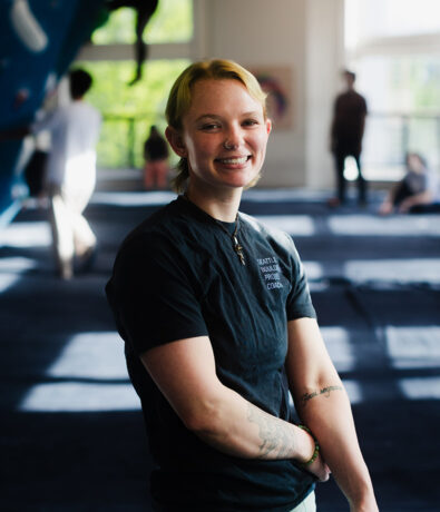 A smiling person with short blonde hair stands indoors, wearing a dark t-shirt. Sunlight streams through large windows in the background, where other people are visible in a blurred, open space in Seattle's personal coaching center.