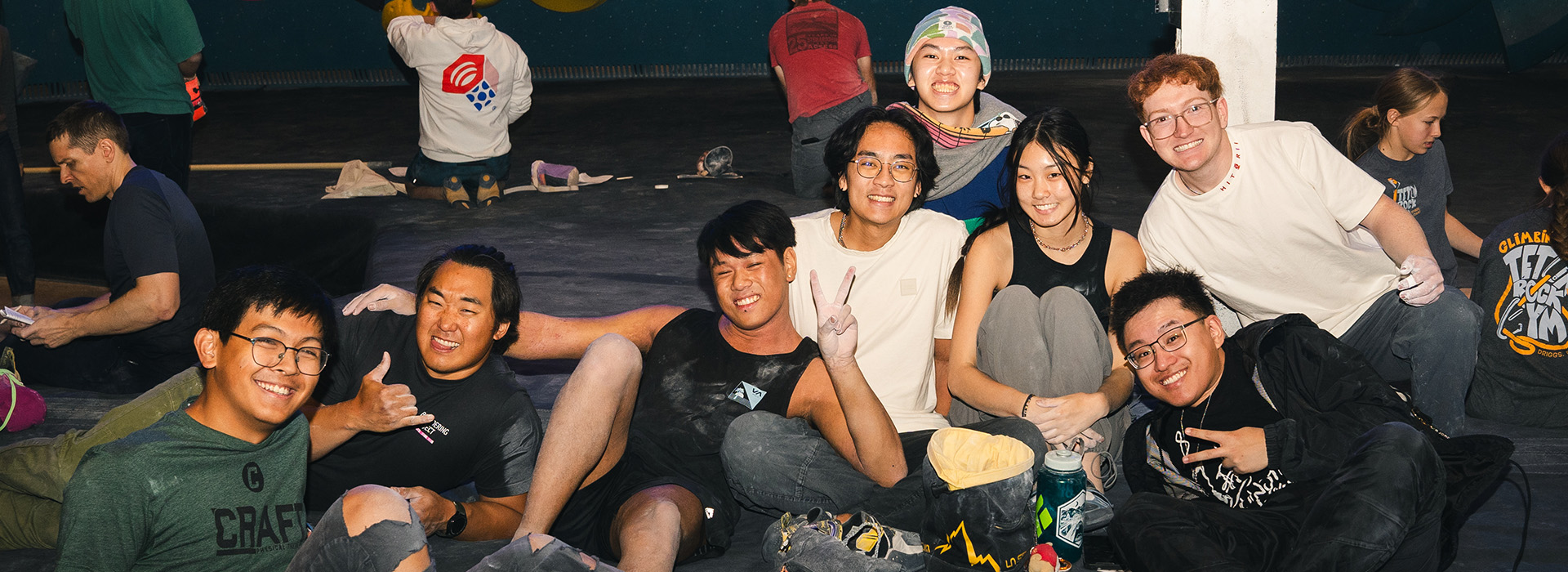 A group of nine young adults sit close together on the floor, smiling and posing for a photo at a Salt Lake City indoor climbing gym. Climbing gear and chalk bags are visible, with waived initiation promos drawing more climbers in the background.