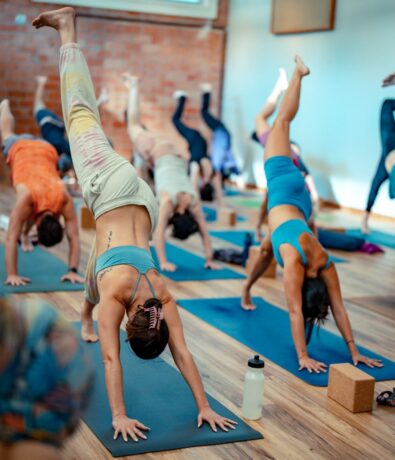 A group of people in colorful workout clothes practice yoga indoors on blue mats in Fremont, each raising one leg in the air. A water bottle and yoga blocks are visible in the foreground near the upper walls.