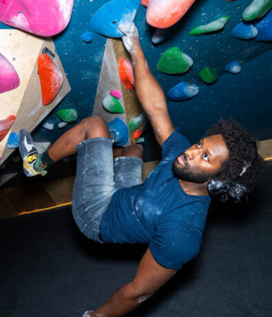 A man with headphones and chalked hands climbs an indoor bouldering wall, confidently gripping colorful climbing holds while wearing a blue shirt, denim shorts, and climbing shoes.