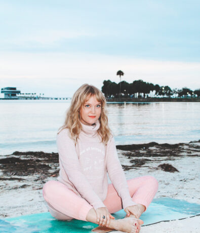 A woman sits cross-legged on a yoga mat on a sandy beach, wearing a light pink sweater and pants. The ocean and distant trees are visible in the background under a cloudy sky.