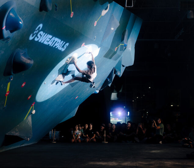 A climber grips an indoor bouldering wall under a spotlight at Boulderfest 2025 in Tempe, with a crowd of spectators watching from below. The wall features large holds and the word SWEATPALS is visible.