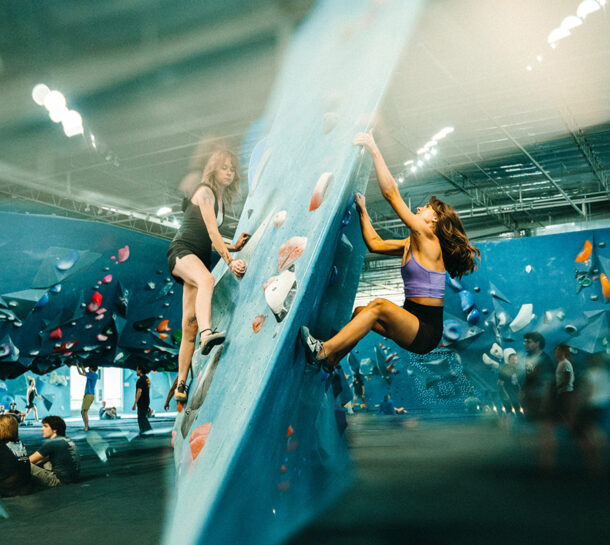 Two women are climbing an indoor bouldering wall with colorful holds, while people watch and relax in the background. The scene is dynamic and filled with natural light.
