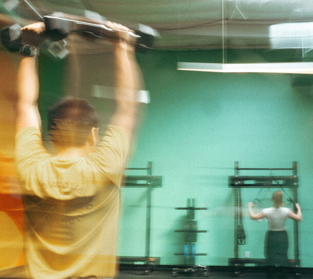 A person in the foreground lifts a dumbbell overhead, while another person in the background uses gym equipment against a green wall; the image is slightly blurred to suggest motion.