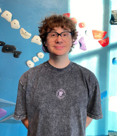 A person with curly brown hair and glasses stands smiling in front of a blue indoor climbing wall with colorful holds in Tempe. They wear a dark, short-sleeved shirt with a circular logo, ready to offer personal coaching.