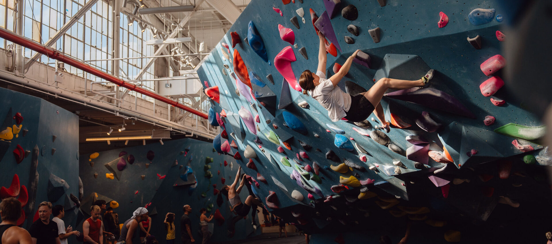A group of people in a bright Brooklyn Climbing Yoga and Fitness gym, with one person scaling a blue climbing wall covered in colorful holds. Others watch or prepare to climb nearby as sunlight streams through large windows above.
