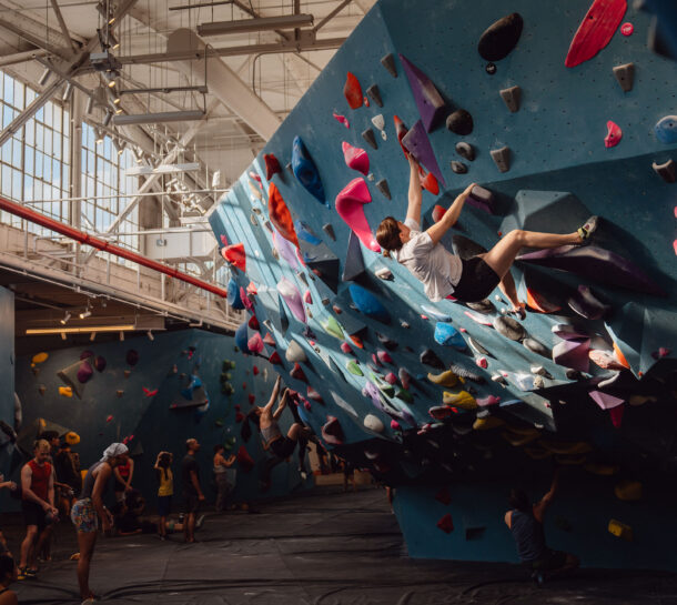 A group of people in a bright Brooklyn Climbing Yoga and Fitness gym, with one person scaling a blue climbing wall covered in colorful holds. Others watch or prepare to climb nearby as sunlight streams through large windows above.
