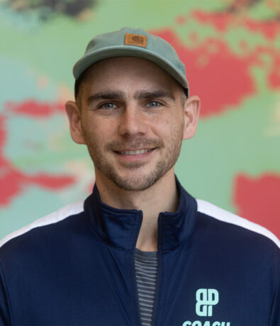 A smiling man wearing a light blue cap and a navy blue jacket with a Coach logo stands in front of a blurry, colorful background, representing the Minneapolis Climb Team.