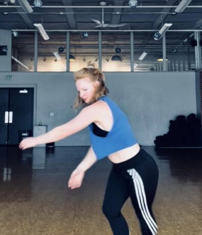 A woman in a blue crop top and black leggings with white stripes dances barefoot in a spacious Fremont studio, mid-movement with arms extended and knees bent, sunlight streaming from the high ceilings and upper walls.