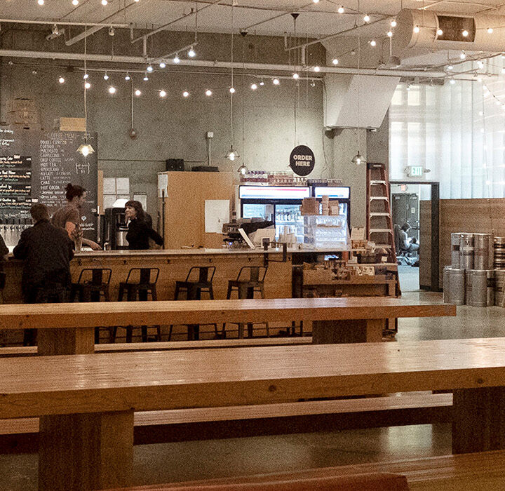 A spacious, modern cafe and bar with wooden benches and tables, string lights overhead, and people working or chatting. The counter and menu are in the background, with a few customers and staff visible at West Wall Cafe.