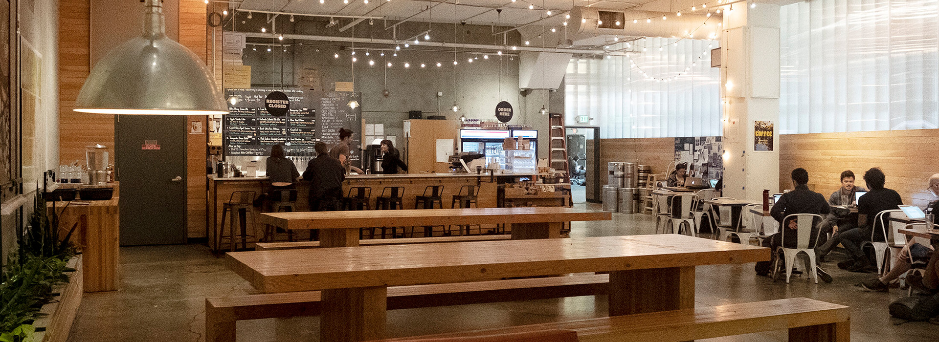 A spacious, modern cafe and bar with wooden benches and tables, string lights overhead, and people working or chatting. The counter and menu are in the background, with a few customers and staff visible at West Wall Cafe.