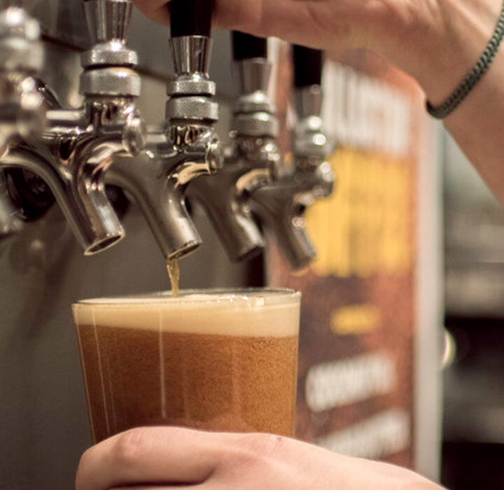 A tattooed person’s hand holds a plastic cup under a row of beer taps at West Wall Cafe, filling it with foaming draft beer. The blurred background hints at the lively ambiance of this popular cafe bar.