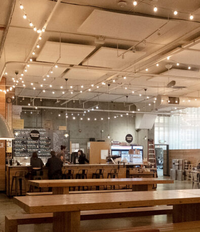 A spacious, modern café with high ceilings, string lights, wooden tables, and a bar counter where a few people are ordering. The space feels open and inviting with a relaxed atmosphere.