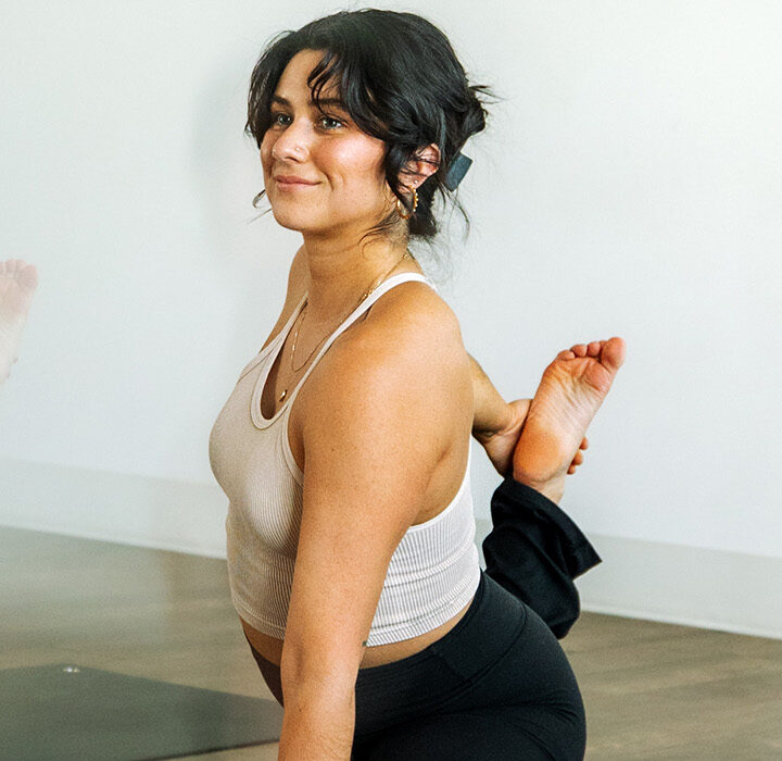 Three women practice yoga indoors, each holding a seated pose with one leg bent back. The central woman looks toward the camera and smiles. Members in Salt Lake City enjoy 24/7 access to serene, softly blurred studios like this one.