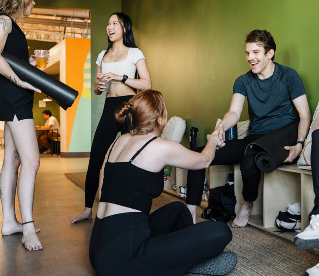 A group of young adults in athletic wear chat and laugh in a yoga studio. One woman shakes hands with a seated man holding a yoga mat, while two others stand nearby, smiling and discussing the benefits of the summer membership.