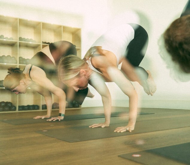 Two women balance on mats in a yoga studio, performing arm-balancing poses, with shelves of weights behind them. The image has a blurred, double-exposure effect—perfect inspiration for your summer membership goals.