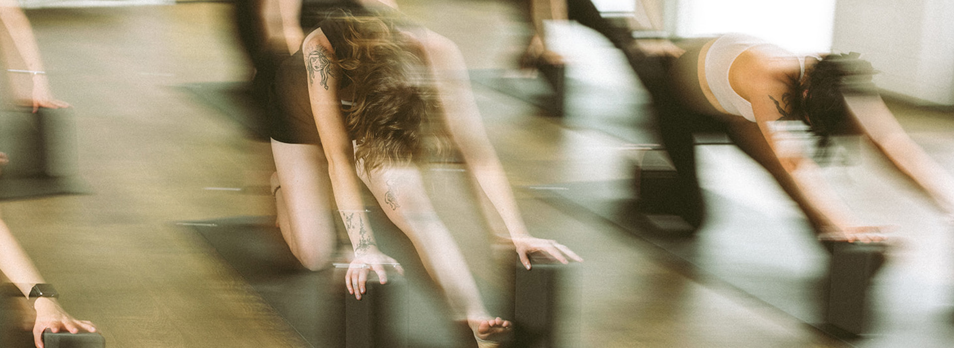 Several people in athletic wear perform yoga stretches on the floor using yoga blocks. The image is softly blurred, emphasizing motion and form on a wooden floor in a bright studio.