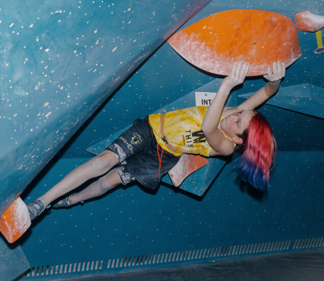 A person with long red and blue hair climbs indoors on an overhanging blue and orange bouldering wall at a Seattle Bouldering Climbing Gym, wearing a yellow tank top and black shorts. They grip large orange holds with both hands and feet.