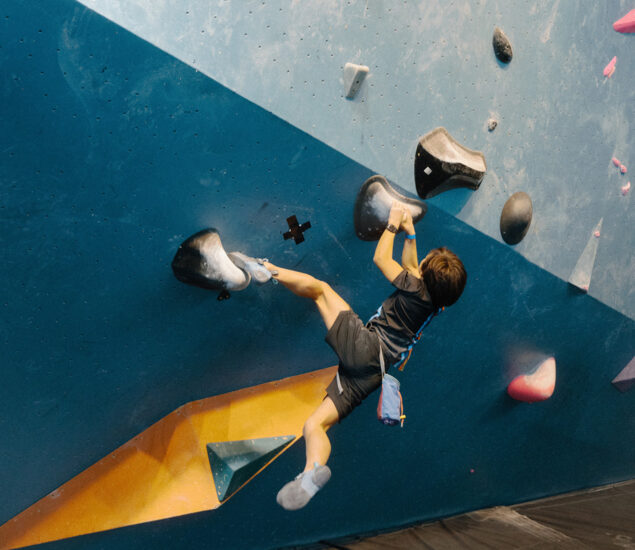 A young boy climbs an indoor bouldering wall at a Seattle climbing gym, gripping large black holds with arms and legs stretched out, wearing a harness, chalk bag, and climbing shoes—perfect for after school clubs.