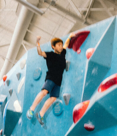 A young boy in a black shirt and blue shorts climbs an indoor rock climbing wall at a Seattle Bouldering Climbing Gym, gripping a red hold with one hand and lifting the other arm. The large, well-lit gym is perfect for After School Clubs.