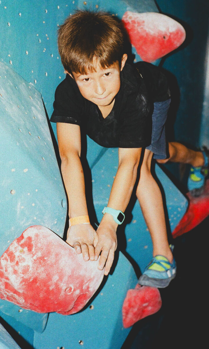 A young boy wearing a black shirt and blue shorts climbs a brightly colored indoor rock wall, gripping large red and white handholds. He looks determined and focused—a great example of the excitement found in our Youth Programs.