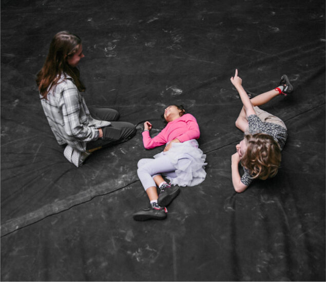 Three children are on a dark floor at Brooklyn Climbing Yoga and Fitness; one sits cross-legged, another lies on their back with eyes closed, and the third lies on their side pointing upward, all appearing relaxed and engaged together.