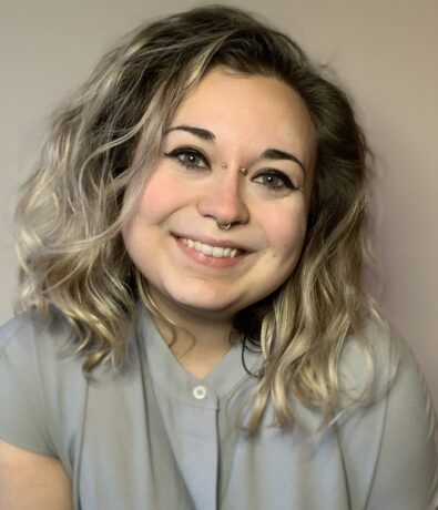 A person with wavy, shoulder-length blonde hair smiling at the camera. They are wearing a light grey shirt and have a nose ring and two nose studs. The background is plain and light-colored.