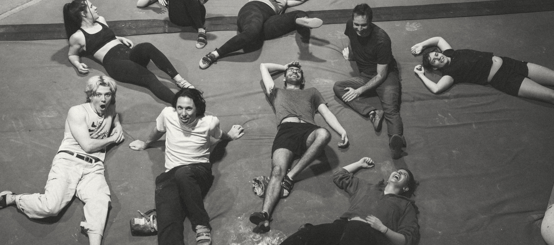 A group of eight people in casual clothes lie and sit relaxed on a padded gym floor, smiling and laughing together. The atmosphere appears joyful and friendly. The image is black and white.