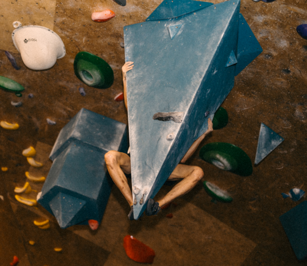 A person is bouldering indoors, mostly hidden behind a large blue triangular climbing hold, with only parts of their arms and legs visible gripping the structure. Colorful climbing holds are attached to the wall.