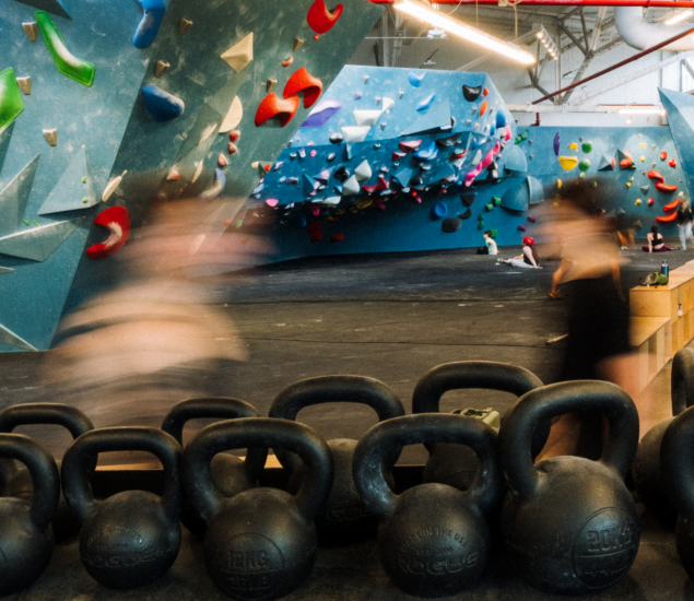 People walk past rows of kettlebells in a brightly lit indoor climbing gym with colorful climbing holds on blue walls in the background. The movement creates a blurred effect.