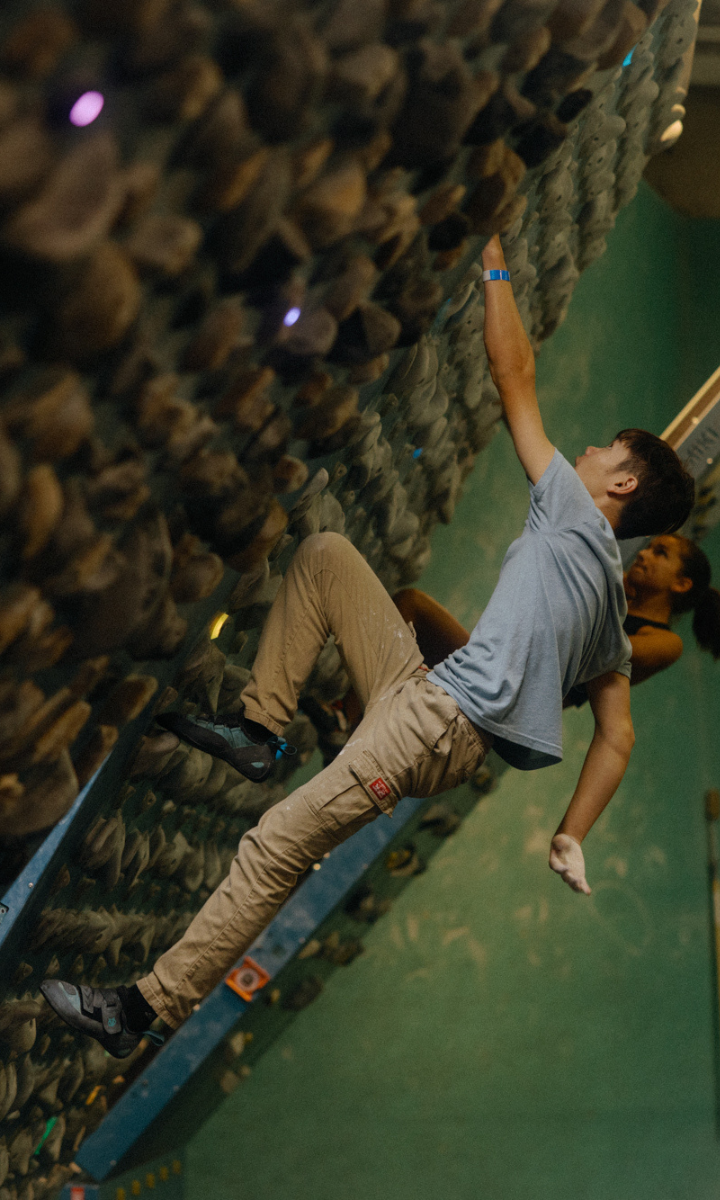 A boy wearing a light blue shirt and beige pants climbs an indoor bouldering wall during the OB/SESSION Board League, reaching for a handhold as another climber follows. The wall is covered in various climbing grips.