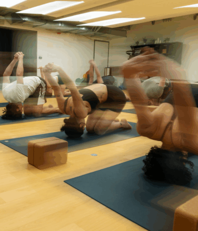 A group of people practice Hot Fusion Yoga indoors on mats, performing a forward bend with arms clasped behind their backs. The motion blur conveys movement and energy in the bright studio, with yoga blocks visible on the floor.