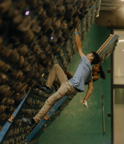 A boy climbs an indoor bouldering wall, reaching up with one hand while gripping holds with his feet. In the brightly lit climbing gym, a climbing training board is visible in the background near another climber.