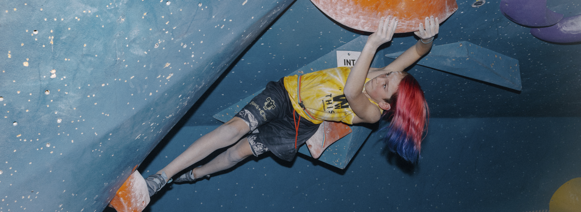 A young person with red and blue hair climbs a steep indoor rock wall, gripping an orange handhold while participating in one of the exciting youth programs Boston has to offer, dressed in a yellow tank top and black shorts.