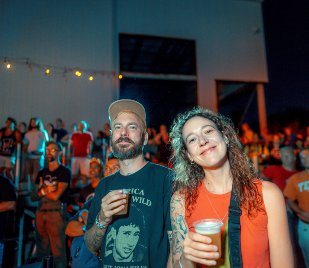 A man and a woman smiling and holding drinks stand in the foreground at an outdoor 2024 Boulderfest event in Austin at night, with a crowd of people behind them under colorful lights.